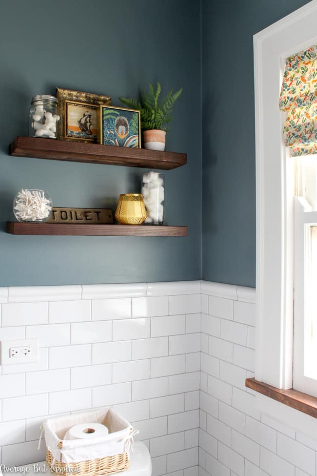 Floating shelves above the toilet are a place to display artwork, useful items for the bathroom like cotton swabs and cotton balls, and a nice candle. Wall color is Charlotte Slate by Benjamin Moore.