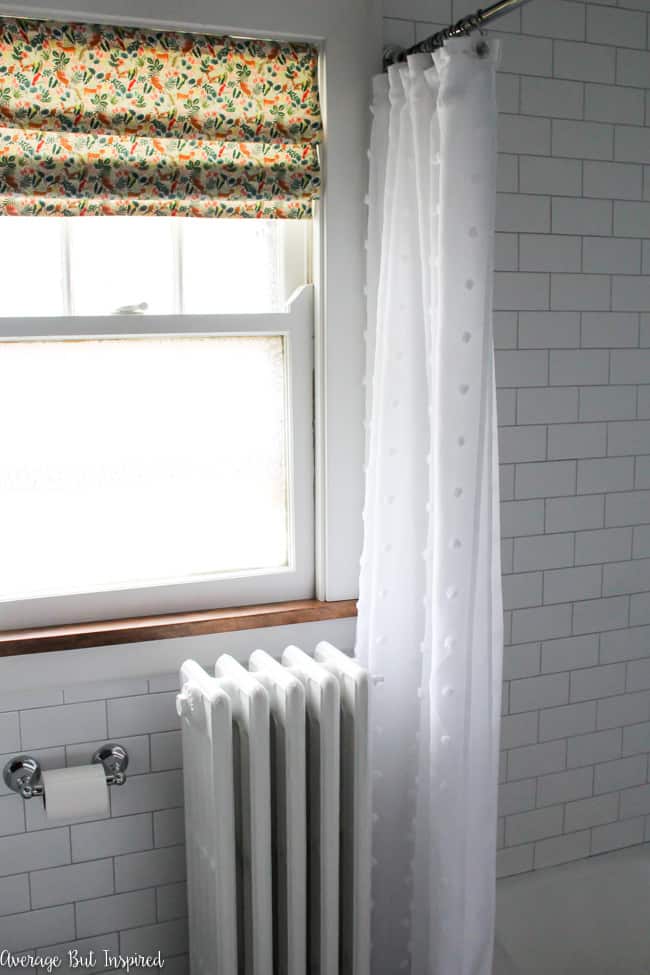 A faux roman shade with pretty fabric and a white shower curtain complement the white subway tile and light gray grout.