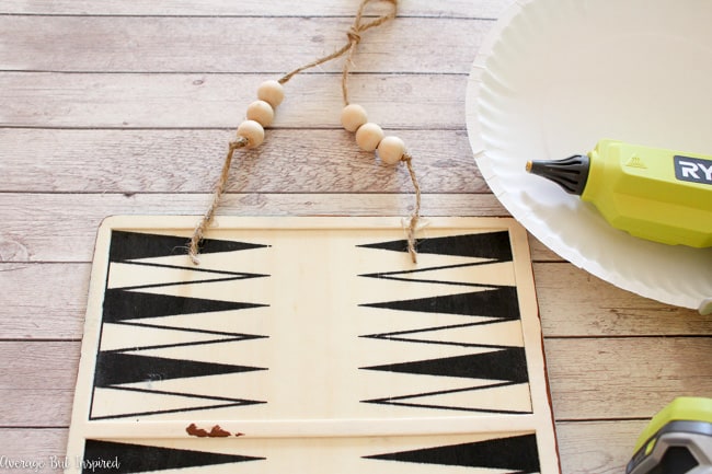 Hot glue the beads and twine to the back of the game board to create a hanger for the vintage tin tile wall art.